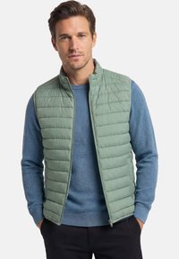 Mann mit kurzen braunen Haaren, der eine hellgrüne gesteppte Weste über einem blauen Pullover und schwarzen Hosen trägt, steht mit den Händen in den Taschen.