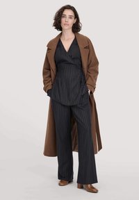 Donna con capelli corti e ricci di colore marrone che indossa un lungo cappotto marrone su una maglietta avvolgente nera a righe e pantaloni larghi abbinati, con stivali marroni.