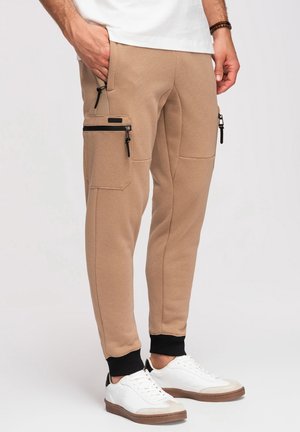 Homme portant un pantalon jogger beige avec des poches zippées noires et des poignets noirs, associé à des baskets blanches avec des semelles marron.