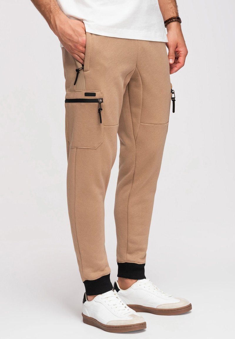 Homme portant un pantalon jogger beige avec des poches zippées noires et des poignets noirs, associé à des baskets blanches avec des semelles marron.