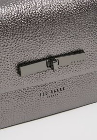 Srebrna torebka kopertówka o teksturowanej powierzchni, gładkim, błyszczącym wykończeniu oraz metalowym zapięciu z czarnym logo "Ted Baker". Widoczne eleganckie detale.