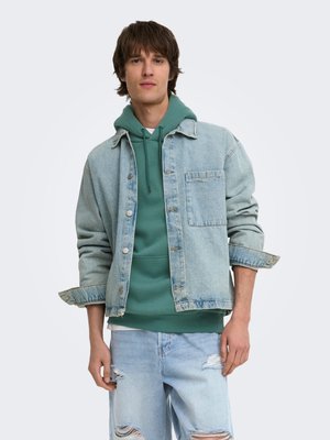 Junger Mann trägt einen grünen Hoodie unter einer hellen Jeansjacke mit hochgekrempelten Ärmeln und zerrissenen hellblauen Jeans, stehend vor einem schlichten Hintergrund.