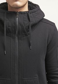 Felpa nera con zip, dotata di cappuccio trapuntato e testurizzato, lacci e tasche frontali. Presenta un rivestimento morbido e polsini a coste.