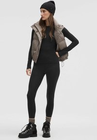 Pehme ja beež vest musta pika varrukaga särgi ja legginsite kohal, koos mustade nööbitavate saapade ja musta kudumisega beanie'ga.