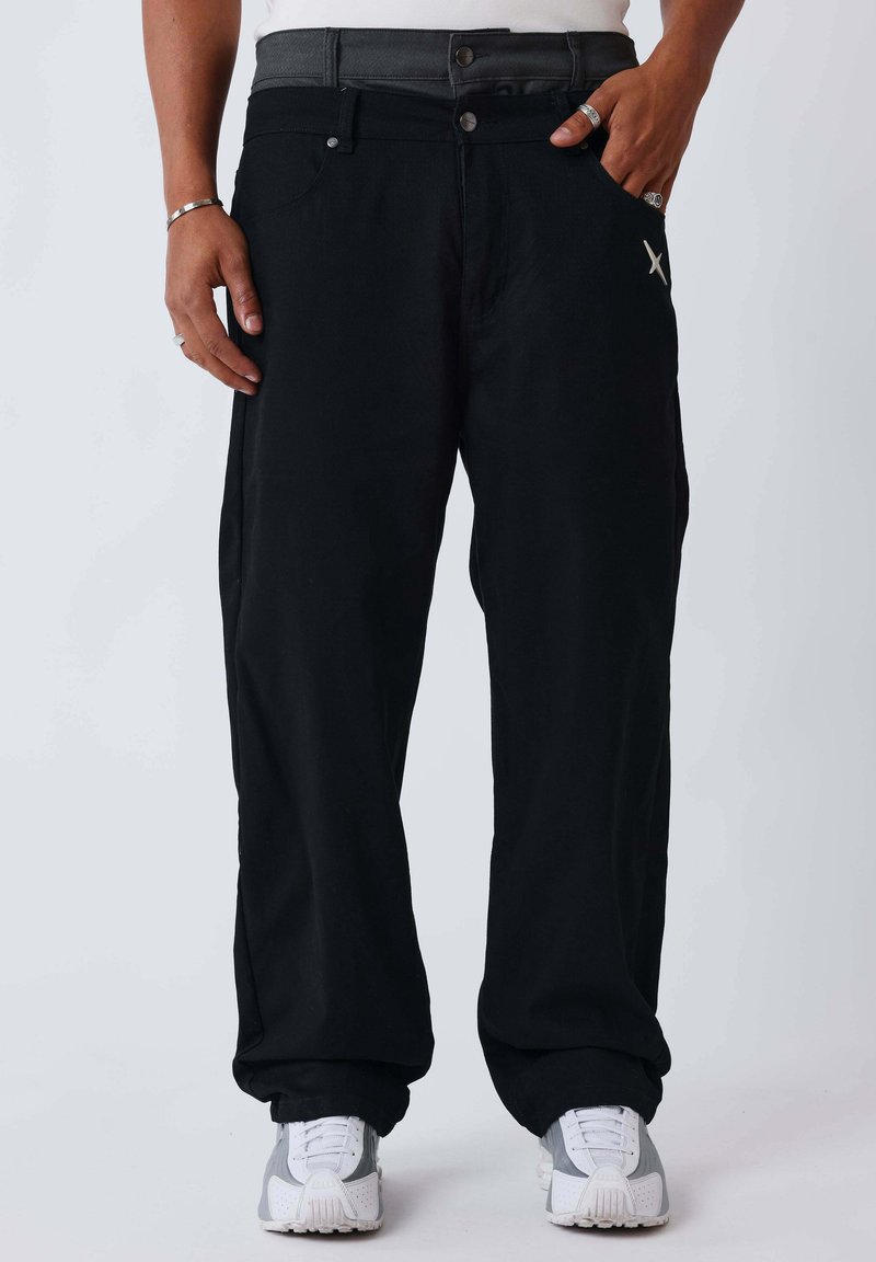 Homme portant un pantalon noir ample avec un petit logo blanc près de la poche, des baskets blanches, des bagues argentées et un bracelet, debout devant un fond uni.