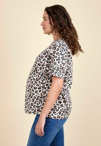 Tričko s krátkým rukávem v leopardím vzoru v béžové barvě s černými a hnědými skvrnami, volný střih. Nosené s modrými džíny, ukazující siluetu z boku.