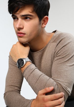Joven con camisa beige de manga larga apoyando la barbilla sobre el puño, lleva un reloj de pulsera plateado con esfera negra en la muñeca izquierda.