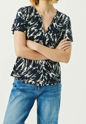 Kurzärmliges Oberteil mit einem marineblauen und weißen abstrakten Muster, einem V-Ausschnitt und einer lockeren Passform, kombiniert mit blauen Jeans.