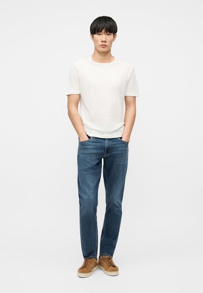 Jeune homme debout, les mains dans les poches, portant une chemise blanche à manches courtes, un jean bleu et des baskets marron sur fond blanc.