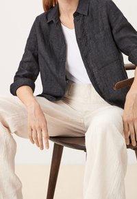 Chemise en lin foncé à boutons avec manches longues et une poche frontale, associée à un pantalon large beige clair, assis sur une chaise en bois.