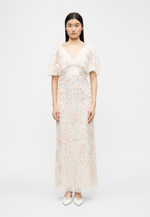 Donna in piedi che indossa un vestito bianco trasparente fino alla caviglia con ricami floreali pastello e maniche corte larghe, abbinato a scarpe bianche con punta affilata.