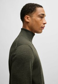 Maglione verde oliva a collo alto con design aderente, tessuto con texture e spalle strutturate. Presenta un collo alto e polsini a coste.