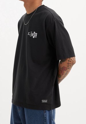 Osoba nosząca czarną koszulkę Levi's z logo na klatce piersiowej i widocznym tatuażem na ramieniu, w połączeniu z niebieskimi dżinsami na jednolitym tle.