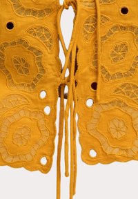Gul broderad textil med blommiga och geometriska utskärningsmönster samt knytband för stängning. Mjukt, texturerat tyg med intrikata detaljer.