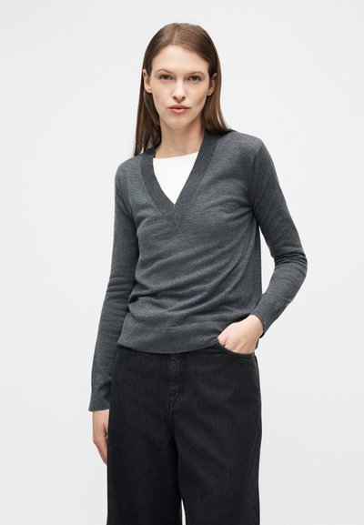 Γυναίκα με ίσια καστανά μαλλιά που φοράει σκούρο γκρι πουλόβερ με V λαιμόκοψη πάνω από λευκό τοπ και μαύρο παντελόνι, στέκεται με το ένα χέρι στην τσέπη.
