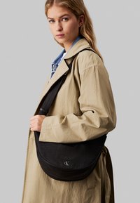 Czarna, teksturowana torba na ramię o zaokrąglonym kształcie z srebrnymi elementami metalowymi. Posiada detal z logo. Modelka nosi beżowy płaszcz z niebieskim kołnierzem.