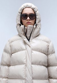 Giacca puffer beige con colletto alto e cappuccio, caratterizzata da una finitura lucida, un design trapuntato e una chiusura con zip. Occhiali da sole scuri completano il look.