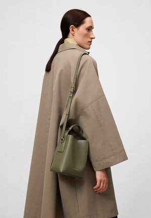 Groene leren emmer tas met een gestructureerd ontwerp, korte handvatten en een verstelbare schouderriem. Draagt door een persoon in een beige jas.