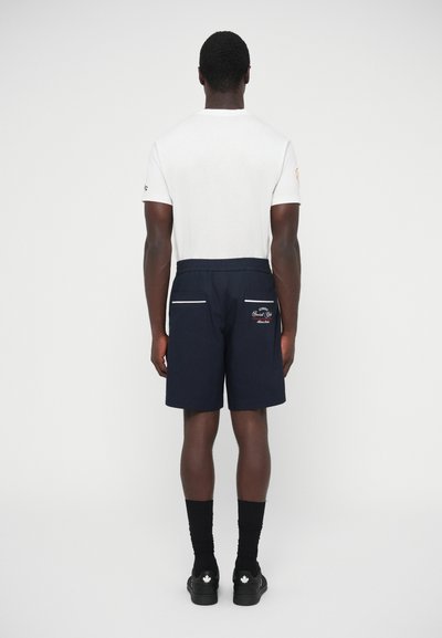 Σορτς σε σκούρο μπλε χρώμα με ελαστική μέση, πλαϊνές τσέπες και λευκές λεπτομέρειες. Συνδυασμένα με εφαρμοστό λευκό μπλουζάκι και μαύρες κάλτσες.