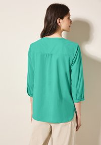Blusa turchese con maniche a tre quarti, scollatura rotonda e dettagli arricciati sul retro. Tessuto leggero, texture liscia, fenditure laterali.