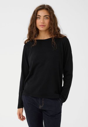 Modèle aux longs cheveux bruns, portant un haut noir uni à manches longues et un jean foncé, sur un fond de studio clair.