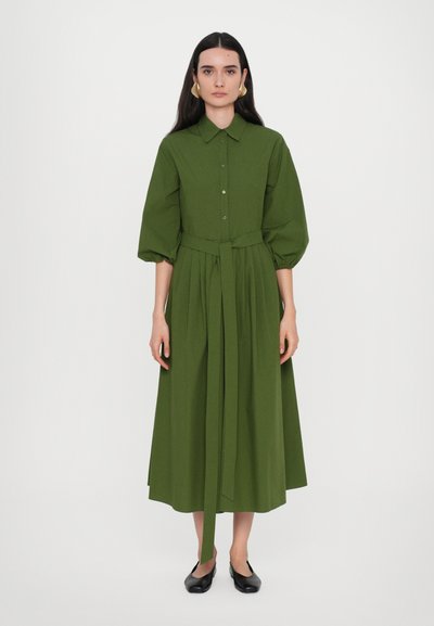 Grünes, knielanges Kleid mit Puffärmeln, tailliertem Gürtel und fließendem Rock. Glatte Textur und knöchellanges Design. Flache schwarze Schuhe getragen.