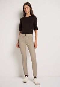 Pantalones ajustados beiges con cintura alta y cierre de doble botón, combinados con una blusa negra de manga corta. El modelo lleva zapatillas blancas.