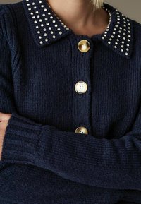 Jūrinis megztinis su raukšlėtu tekstūra, aptrauktas apykakle, papuošta skaidriais kalnų krištolais ir auksinėmis mygtukais priekyje.