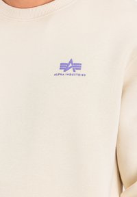 Beige sweatshirt med ribbet rund hals og et lille lilla logo med teksten "Alpha Industries" på venstre brystområde.