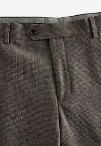 Braune strukturierte Hose mit flacher Vorderseite, ausgestattet mit einem einzelnen Knopfverschluss und einem gemusterten Innenbund für zusätzliche Details.