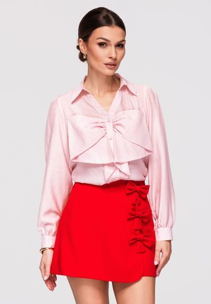 Femme portant une blouse rose pâle avec un grand nœud et une jupe rouge vif ornée de trois nœuds sur les côtés, debout devant un fond uni.
