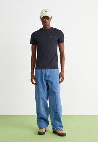 Tricou scurt cu mânecă, de culoare bleumarin, cu un buzunar, asortat cu pantaloni de denim supradimensionați, de culoare albastru deschis, și pantofi bej. Persoana poartă o șapcă bej.