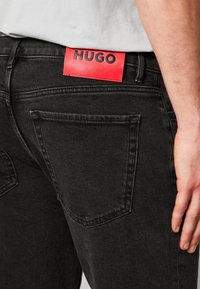 Musta farkkujen materiaali, jossa on punainen nahkainen tagi, jossa lukee "HUGO." Suunnittelussa on kaksi taka-taskua ja sileä pinta.