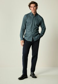 Grünes gemustertes Langarm-Hemd mit Knopfleiste, das mit einem klassischen Kragen und Manschetten versehen ist, kombiniert mit dunklen Hosen und schwarzen Schuhen.