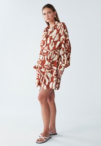 Brązowo-kremowa sukienka koszulowa w wzory z paskiem, długimi rękawami i guzikami, noszona z białymi płaskimi sandałami. Gładka faktura materiału.