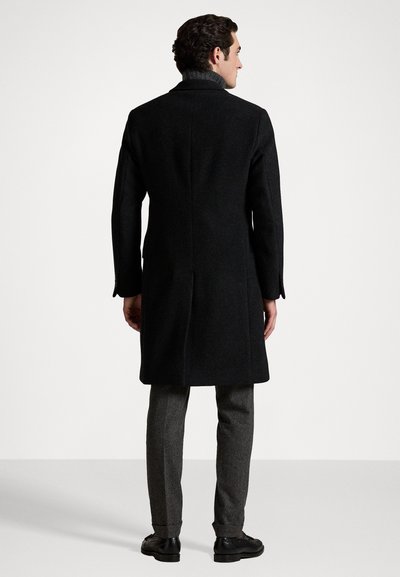 Black wool coat with a tailored fit, a high collar, single vent, and long sleeves, paired with gray textured trousers and black shoes.