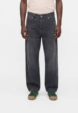 Homme portant un jean noir ample et des baskets vertes, debout devant un fond uni, mains relâchées le long du corps.
