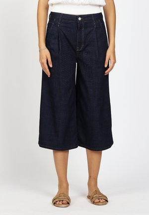 Frau trägt dunkelblaue, weit geschnittene, gekürzte Jeanshosen und bequeme, beige Sandalen mit offenem Zehenbereich, steht vor einem schlichten weißen Hintergrund.