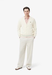 Vit cardigan över ljusrosa polo, i kombination med krämfärgade byxor och beige sneakers. Har gröna randiga accenter vid cardiganens nederkant.