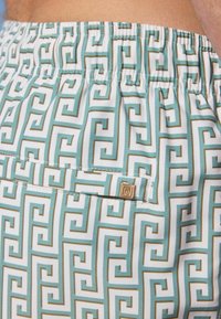 Geometrikus mintázattal ellátott úszóshorts, teal és fehér színben, arany részletekkel és elasztikus derékkal, könnyű anyagból készült.