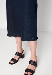 Marinblå midi-klänning med subtilt texturerat mönster, kombinerad med svarta läderklackar med blockklack och breda remmar samt öppen tå.