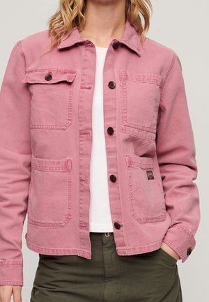 Femme portant une veste rose à boutons avec quatre poches avant sur une chemise blanche et un pantalon vert olive.
