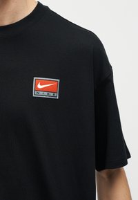 Κοντινό πλάνο ενός ατόμου που φοράει μαύρο μπλουζάκι με ένα μικρό ορθογώνιο κόκκινο και γκρι λογότυπο Nike στο στήθος.