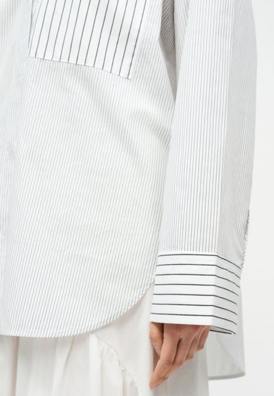 Κοντινό πλάνο ενός λευκού πουκαμίσου με μακριά μανίκια και λεπτές κάθετες ρίγες, με μανσέτα με πιο φαρδιές ρίγες στον καρπό, που φοριέται πάνω από μια λευκή φούστα.