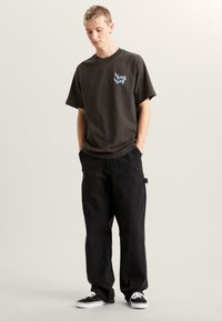 Zwart katoenen T-shirt met een blauwe grafische print op de linkerborst, gecombineerd met losse zwarte broek en zwarte canvas sneakers.