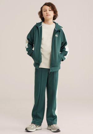 Bambino con tuta verde e bianca, mani nelle tasche della giacca, in piedi con sicurezza su uno sfondo neutro.