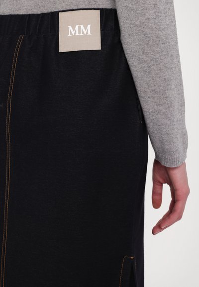 Farmernadrág szoknya narancssárga varrással és oldalsó hasítékokkal, a négyzet alakú címkén fehér "MM" felirattal. Részben látható szürke pulóver ujja.