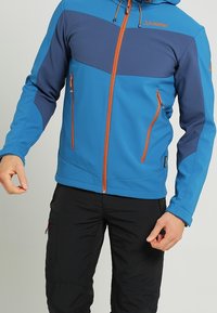 Blauwe softshell jas met marineblauwe accenten, oranje ritsdetails en een capuchon. Inclusief twee ritszakken aan de voorkant.