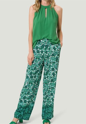 Top verde senza maniche con dettaglio a occhiello; pantaloni verdi larghi e decorati con motivi floreali; sandali verdi coordinati.