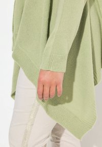 Ljusgrön stickad tröja med en lös, asymmetrisk design, med ribbade fållar och en mjuk textur, ihop med off-white byxor.